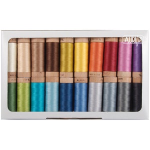 Pode incluir: Uma caixa com 18 carretéis de fio Aurifil em uma variedade de cores. Os carretéis estão dispostos em duas filas de nove. Os carretéis são de madeira e têm um rótulo branco com o logotipo Aurifil. O fio é feito na Itália.