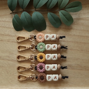 May include: Five keychains with gold metal rings and black cords. Each keychain has a wooden bead, a white bead with the letter "P" and a white bead with the letter "A". Each keychain also has a colorful flower charm. The flower charms are peach, green, yellow, pink, and brown.