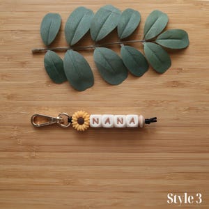 May include: A keychain with the word "NANA" spelled out in white letter beads. A small yellow sunflower charm and a silver clasp are attached. The keychain is on a wooden surface with green leaves.