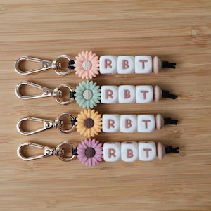 May include: Four keychains featuring a gold-tone metal lobster clasp, a silicone sunflower charm in peach, mint, yellow, or mauve, and three white square beads with the brown letters R, B, and T, finished with a small wooden bead on a black cord.