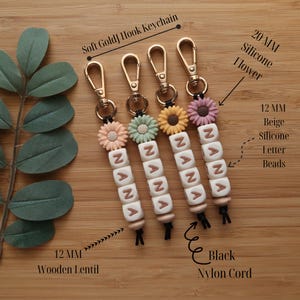 May include: Four keychains with soft gold hooks, each featuring a 20mm silicone flower in different colors. The keychains have beige silicone letter beads spelling "NANA" and are finished with a 12mm wooden lentil and black nylon cord.