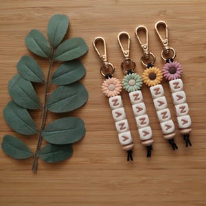 May include: Four keychains with gold-colored clasps, each featuring a daisy flower in different pastel colors. Below the flowers are white letter beads spelling "NANA". The keychains are on a wooden surface with a green leafy branch.