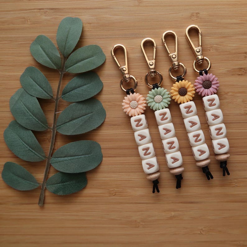 May include: Four keychains with gold-colored clasps, each featuring a daisy flower in different pastel colors. Below the flowers are white letter beads spelling "NANA". The keychains are on a wooden surface with a green leafy branch.