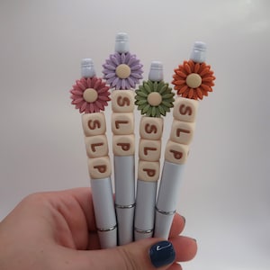 May include: Four white pens with decorative flower tops in shades of pink, purple, green, and orange. Each pen has the letters "SLP" on a series of small, square beads. The pens are held in a hand.