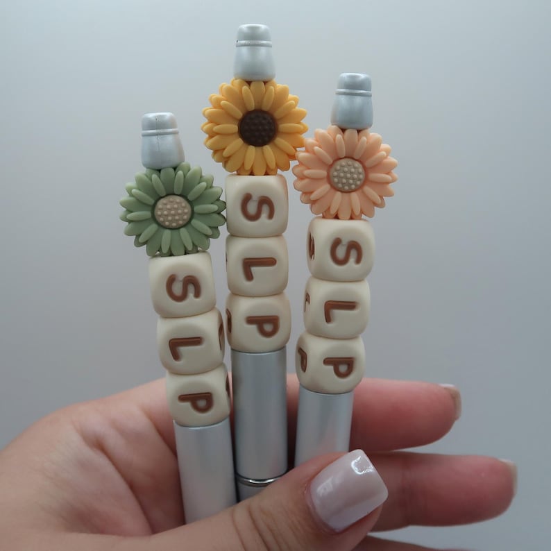 May include: Three silver pens with white beads spelling "SLP" and topped with a sunflower, a peach flower, and a green flower.