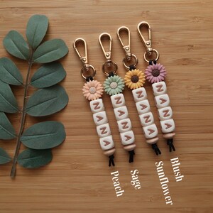 May include: Four keychains with gold-colored clasps, each featuring the name "NANA" spelled out in white letter beads. Each keychain has a different colored flower and the text "Peach", "Sage", "Sunflower", and "Blush".