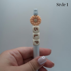 May include: A white pen with a peach-colored flower charm and white letter beads spelling "SLIP".