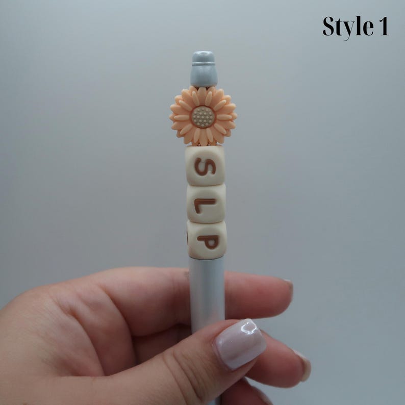 May include: A white pen with a peach-colored flower charm and white letter beads spelling "SLIP".