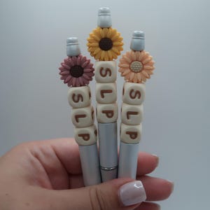 May include: Three silver pens with white and brown bead accents spelling "SLIP". Each pen is topped with a different colored flower: yellow, peach, and pink.