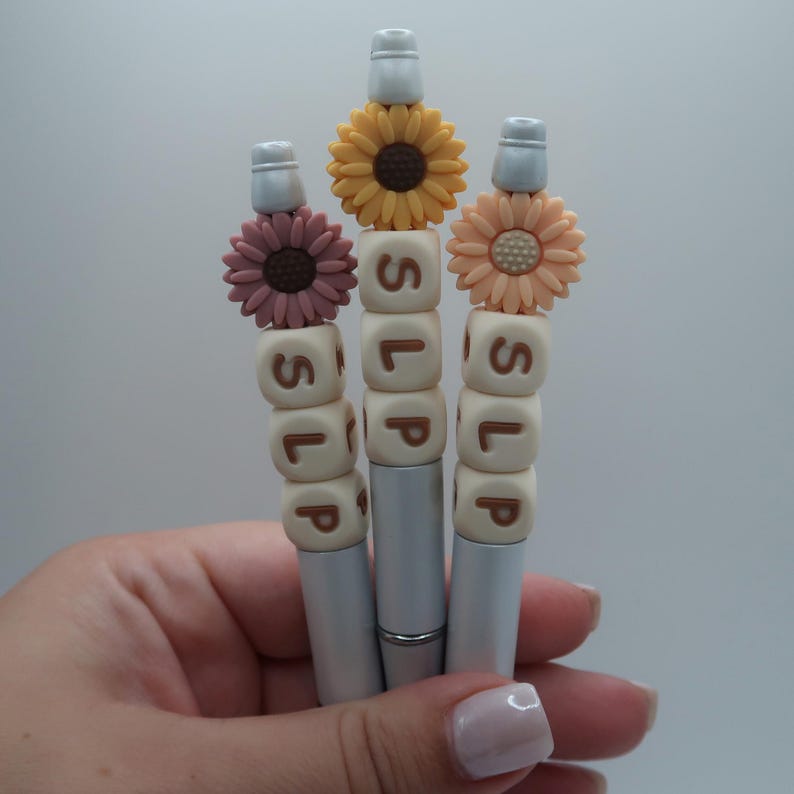 May include: Three silver pens with white and brown bead accents spelling "SLIP". Each pen is topped with a different colored flower: yellow, peach, and pink.