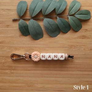 May include: A keychain with the name "NANA" spelled out in white letter beads. A peach-colored flower and a gold-colored clasp are attached. The keychain is displayed on a wooden surface with green leaves.