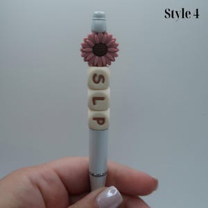 May include: A silver pen with a white top and a pink flower charm. The pen has four white beads with the letters "SLP" spelled out in brown.