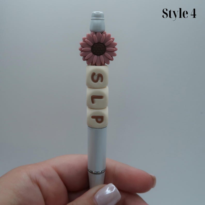 May include: A silver pen with a white top and a pink flower charm. The pen has four white beads with the letters "SLP" spelled out in brown.