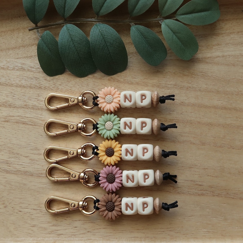 Puede incluir: Cinco llaveros con anillas divididas doradas, cuerda negra, cuentas de madera y cuentas de letras blancas que deletrean "NP". Cada llavero presenta un dije de flor de color diferente: amarillo, verde, naranja, rosa y marr&oacute;n.
