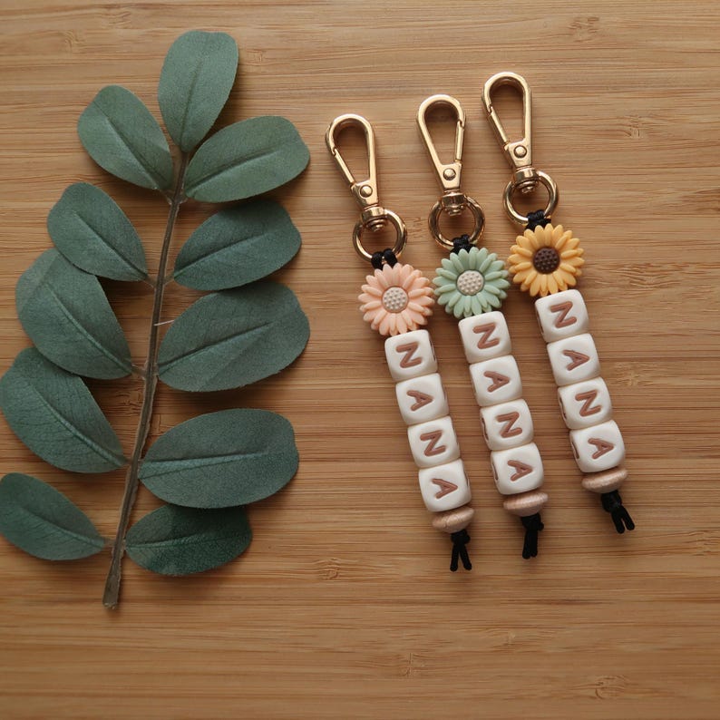 May include: Three keychains with gold-colored clasps and the name "NANA" spelled out in white letter beads. Each keychain features a different colored flower: peach, mint green, and yellow. The keychains are on a wooden surface.