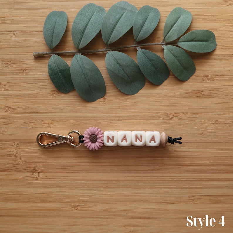 May include: A keychain with the name "NANA" spelled out in white letter beads. A pink flower and gold-colored clasp are attached. The keychain is on a wooden surface with green leaves.