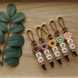 Puede incluir: Cinco llaveros dorados con dijes de girasol y cuentas de letras que deletrean "NP". Cada llavero tiene un dije de girasol de un color diferente: marr&oacute;n, rosa, amarillo, verde y blanco.