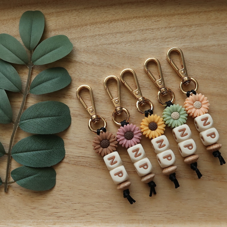 Puede incluir: Cinco llaveros dorados con dijes de girasol y cuentas de letras que deletrean "NP". Cada llavero tiene un dije de girasol de un color diferente: marr&oacute;n, rosa, amarillo, verde y blanco.