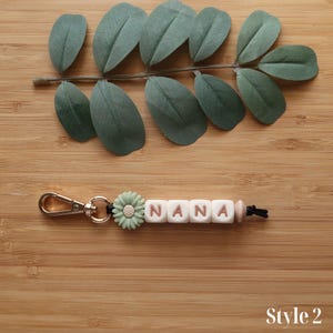 May include: A keychain with the word "NANA" spelled out in white letter beads with brown letters. A light green flower bead and a gold-colored clasp are attached. The keychain is on a wooden surface with green leaves.