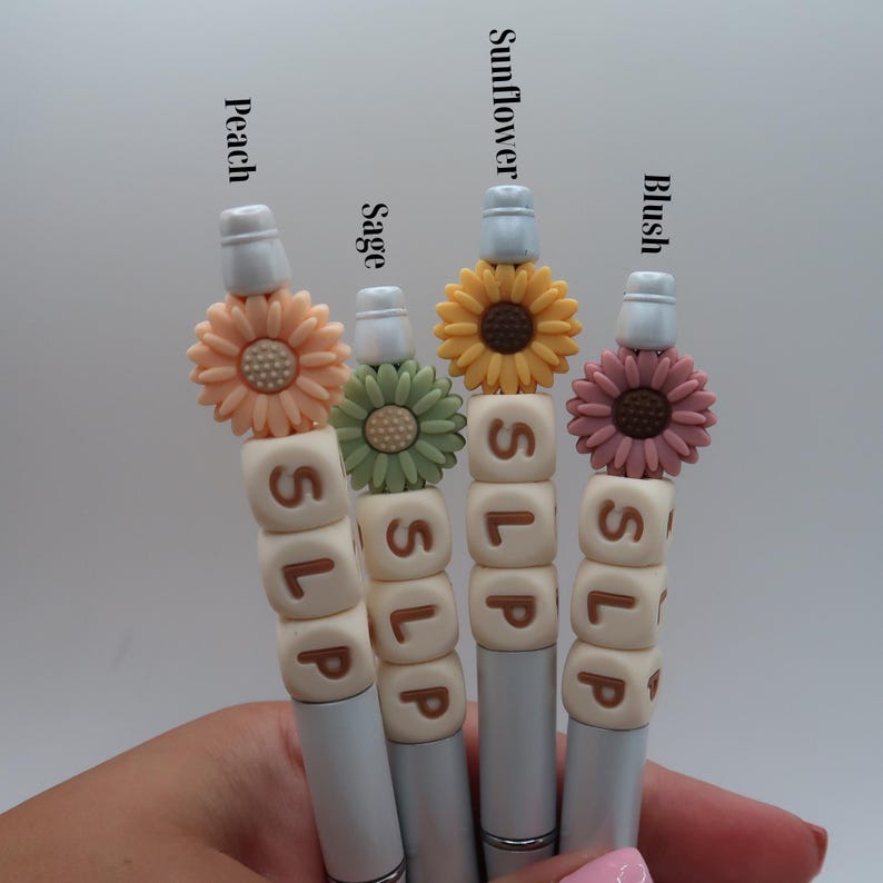 May include: Four white pens with different colored flower charms and the word "SLIP" spelled out in brown letters on the pen barrels. The flower charms are peach, yellow, green, and pink.