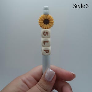 May include: A white pen with a sunflower charm and the word "SLP" spelled out in brown letters on white blocks.