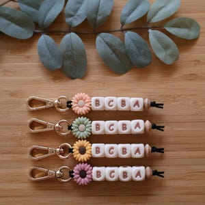 May include: Four keychains with wooden beads, a gold metal ring, and a silicone flower charm. Each keychain has the letters "BCBA" spelled out on white silicone beads.