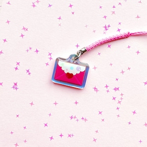 May include: A pink acrylic charm shaped like a sealed envelope with a scalloped white flap, a small red heart, and light blue circular accents. The charm is attached to a braided pink cord, featuring a glossy finish and a clear border.