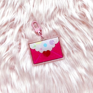 May include: A pink acrylic keychain featuring a letter design with a red heart and colorful accents. The keychain has a pink clasp and is set against a fluffy, pink background.