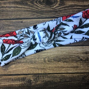 May include: White knotted headband with a floral print featuring red roses, green leaves, and the text "Cosmic" and "Mama".