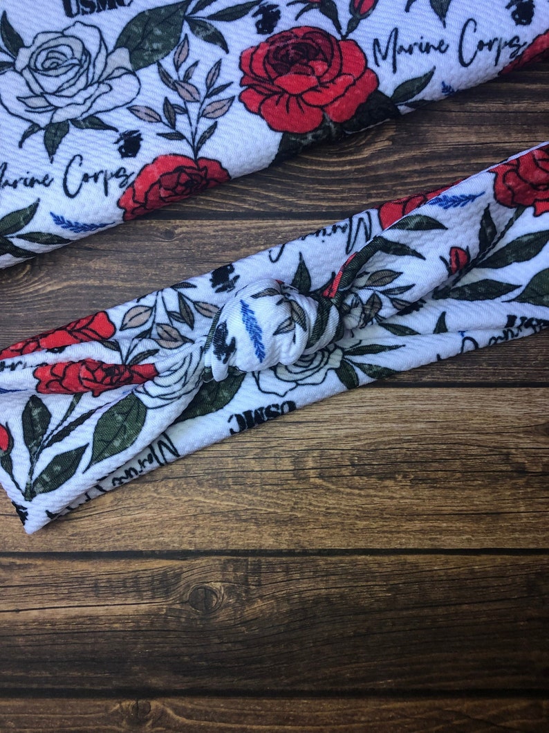 May include: White headband with a knotted design and a floral print featuring red roses, green leaves, and the text "USMC" and "Marine Corps".