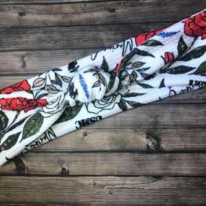 May include: White fabric headband with a floral pattern featuring red roses, green leaves, and white flowers. The headband is tied in a knot and has a textured surface.
