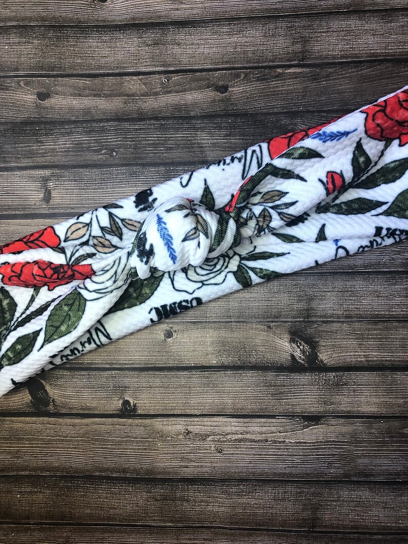 May include: White fabric headband with a floral pattern featuring red roses, green leaves, and white flowers. The headband is tied in a knot and has a textured surface.