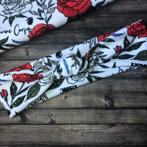 May include: White headband with a floral pattern featuring red roses, green leaves, and the text "Marine Corps".