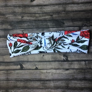 May include: White knotted headband with a floral print featuring red roses, white flowers, and green leaves. The headband has the words "Cosmic" and "Magic" printed on it.