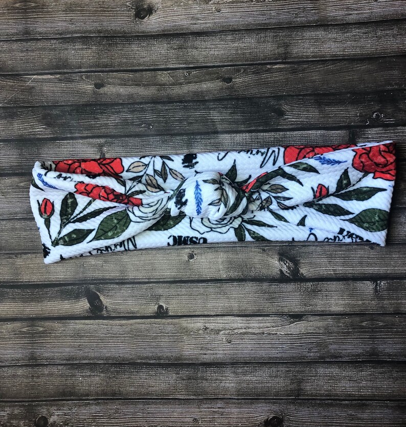 May include: White knotted headband with a floral print featuring red roses, white flowers, and green leaves. The headband has the words "Cosmic" and "Magic" printed on it.