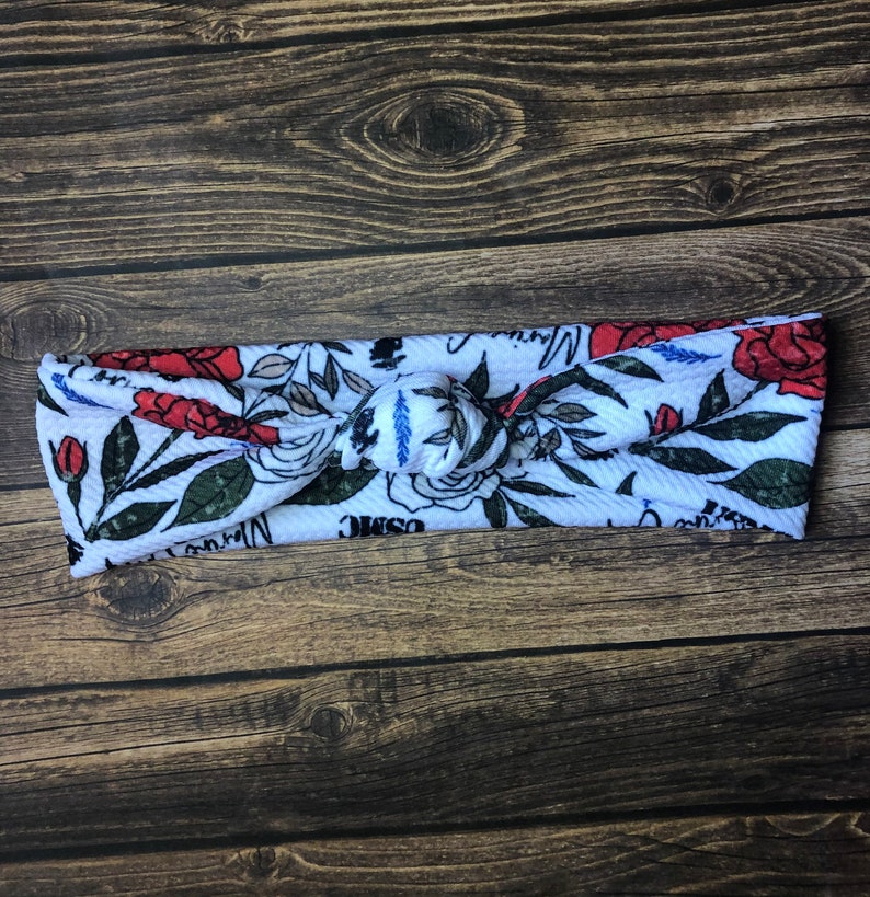 May include: White knotted headband with a floral pattern featuring red roses, green leaves, and the words "Cosmic Warrior".