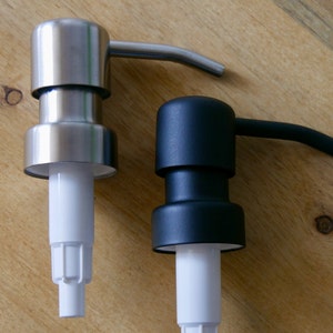 Könnte beinhalten: Zwei Seifenspender, einer silberfarben und einer schwarz, mit weißen Pumpköpfen. Beide Spender haben einen gebogenen Auslauf.