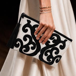 Puede incluir: Clutch rectangular con un diseño en blanco y negro. El clutch presenta una borla negra y un patrón de remolino. El clutch lo sostiene una persona que lleva pulseras doradas y un vestido blanco.