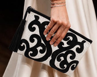 May include: A rectangular clutch bag with a black and white design. The clutch features a black tassel and a swirling pattern. The bag is held by a person wearing gold bracelets and a white dress.