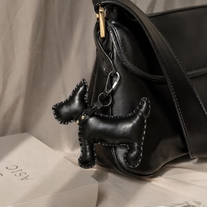Puede incluir: Un bolso de hombro de cuero negro con un colgante en forma de perro sujeto a un cierre de metal dorado. El colgante está hecho de cuero negro con costuras grises visibles en los bordes y un detalle de collar con tachuelas doradas.