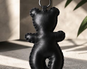 May include: A black leather bear-shaped keyring featuring visible stitched edges and a matte finish. The bear is attached to a black metal ring and a sturdy black metal lobster clasp, providing a secure way to attach it to bags or keys.