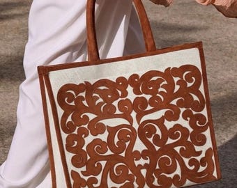 May include: A rectangular tote bag with a brown suede handle and trim. The bag features a cream-coloured background with a brown, swirling, ornate design. The bag is carried by a person wearing white trousers and a peach-coloured top.