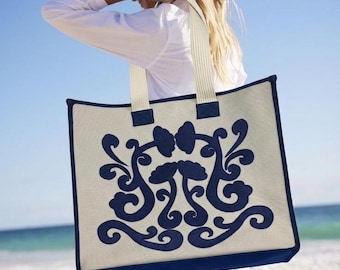 May include: A rectangular tote bag with a cream-coloured background and a navy blue decorative design. The bag has navy blue trim and cream-coloured straps. The bag is being held by a person on a beach.