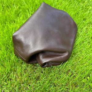 Puede incluir: Un bolso de cuero marr&oacute;n oscuro con una silueta suave, desestructurada y plegada. El art&iacute;culo presenta un acabado liso y mate, con una forma redondeada y flexible que parece maleable, destacando la textura natural del cuero marr&oacute;n oscuro.