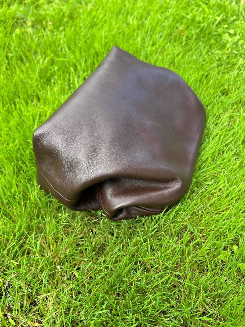 Puede incluir: Un bolso de cuero marr&oacute;n oscuro con una silueta suave, desestructurada y plegada. El art&iacute;culo presenta un acabado liso y mate, con una forma redondeada y flexible que parece maleable, destacando la textura natural del cuero marr&oacute;n oscuro.