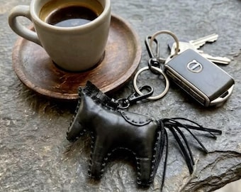May include: A black leather horse-shaped keyring featuring visible stitching along the edges and a fringe tail, attached to a metal ring with a black car key fob. The keyring is positioned next to a ceramic cup filled with dark coffee.