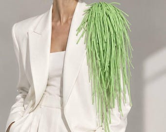 May include: A crisp white blazer featuring a prominent, long, light green fringe attachment on the left shoulder. The blazer has a structured lapel, a single-breasted design, and is paired with matching white trousers and a white top.