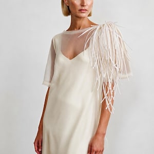 Puede incluir: Un vestido sin mangas en color blanco roto con una capa de tul transparente y escote en V. El hombro izquierdo está adornado con un grupo de tiras de tela blanca largas y finas que cuelgan, creando una apariencia texturizada y en capas.