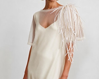 May include: An off-white sleeveless dress featuring a sheer tulle overlay with a V-neckline. The left shoulder is adorned with a cluster of long, thin, white fabric fringe strips that drape downwards, creating a textured and layered appearance.