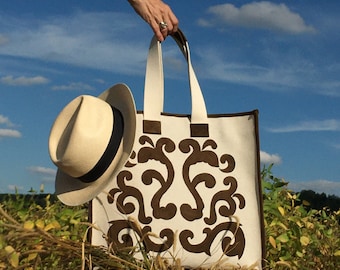 May include: A white canvas tote bag with brown suede embroidery of a floral design. The bag has two handles and is being held by a hand.